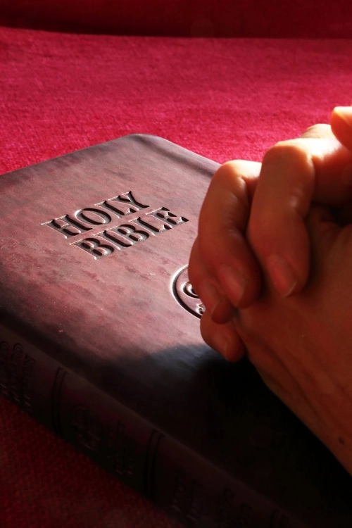 Someone holding hands in prayer posture overlooking a closed Bible on a red surface