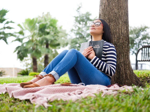 A pretty young woman sitting next to a tree and holding a bible close to her chest to depict reading the Bible conversationally