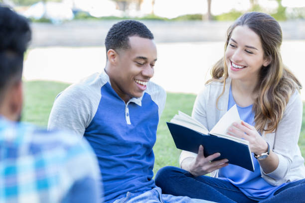 College students relax outdoors on campus while reading from a book to depict community .